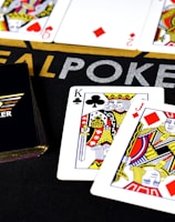 Poker-themed setup featuring a deck of cards on a table with the words 'REAL POKER' visible. Two cards, the King of Clubs and Jack of Diamonds, are prominently displayed. The design is vibrant, with detailed illustrations on the cards.