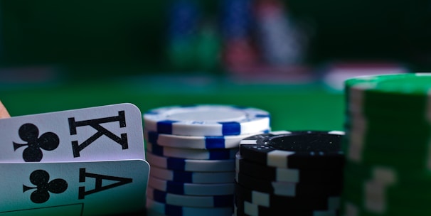 A casino scene featuring two playing cards, the ace and king of clubs, partially concealed by a player's hand. Nearby, there are stacks of multicolored poker chips on a green felt table, with blurred colorful background elements suggesting other chip stacks or cards.