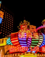 Bright neon lights and colorful signage adorn the exterior of Casino Lisboa, creating a vibrant and lively atmosphere. The façade features bold, multicolored designs with a mix of English and Chinese characters prominently displayed. The lights form intricate patterns and shapes, contributing to the energetic ambiance of the scene.