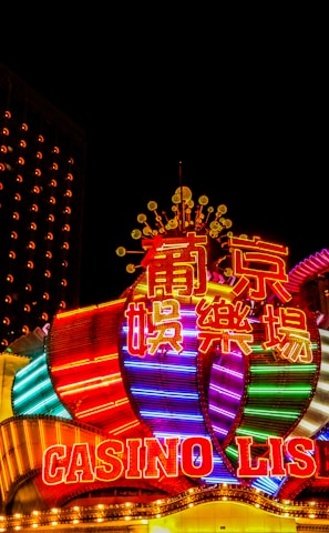Bright neon lights and colorful signage adorn the exterior of Casino Lisboa, creating a vibrant and lively atmosphere. The façade features bold, multicolored designs with a mix of English and Chinese characters prominently displayed. The lights form intricate patterns and shapes, contributing to the energetic ambiance of the scene.