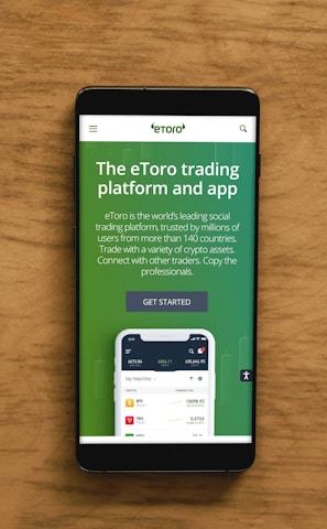 A smartphone displaying an advertisement for an online trading platform called eToro. The screen shows a green background with text promoting the platform as a leading social trading platform. A button labeled 'Get Started' is visible, along with a partial view of a mobile app screen below.