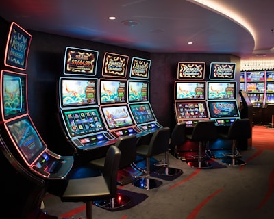 A row of brightly lit slot machines in a casino setting, each with colorful displays and various game themes. The machines are lined up against a dark wall and are unoccupied, with cushioned chairs placed in front of them. The area is dimly lit except for the glow from the screens, creating a vibrant and engaging atmosphere.