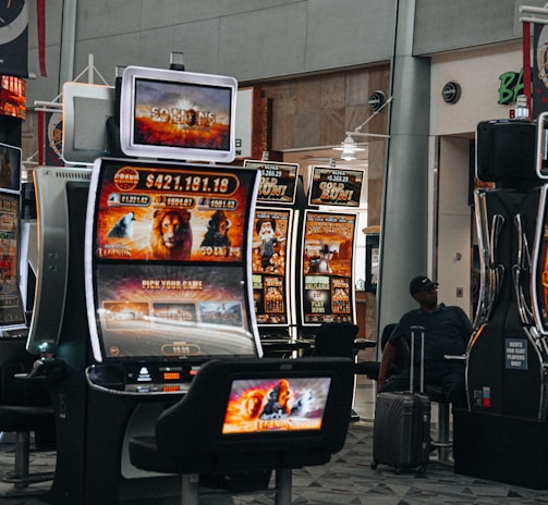 A collection of slot machines is situated inside a building with bright, colorful screens displaying various games and gaming themes. A person is seated nearby, seemingly relaxed with luggage beside them, and the atmosphere is lively with electronic displays.
